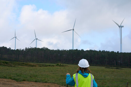 Rear View Of Female Worker With A White Helmet And Yellow Vest In Front Of Wind Turbines In A Rural Renewable Energy Area