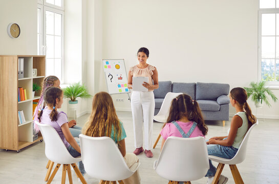 Friendly young woman conducting psychological training for a group of elementary school children girls sitting in a circle in the classroom and listening their psychologist speaker.