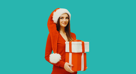 Christmas portrait of happy smiling woman holding gift box in her hands wearing red santa claus hat on blue studio background