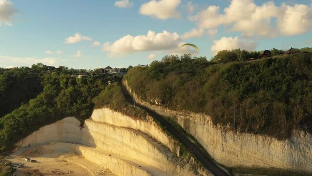 Drone view of a man flying on a yellow paraglider over a hill and trees towards the sea waves near the rocks. Active paragliding flight over seascape with clear sky in Bali. Extreme sport