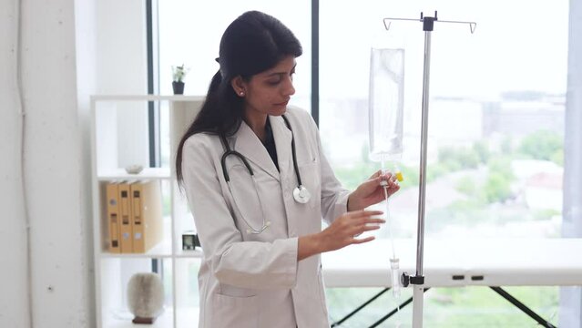 Adult female doctor in white lab coat standing near medical dropper and controlling liquid medication. Indian woman preparing infusion drip for intravenous treatment system.