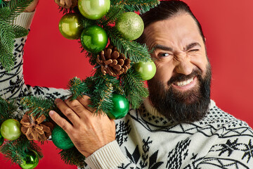 happy bearded man in Christmas sweater holding decorated wreath with baubles on red backdrop