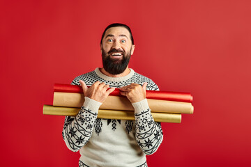 happy bearded man in Christmas sweater holding wrapping paper on red backdrop, holiday sales