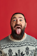 happy bearded man in sweater with ornament sticking tongue out on red backdrop, Christmas
