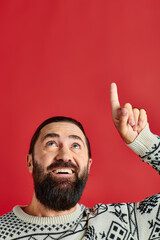 happy bearded man in Christmas sweater with ornament pointing up on red backdrop, winter holidays