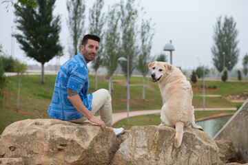 Handsome young man with beard and his large dog sitting on rocks looking back. The dog is a brown...