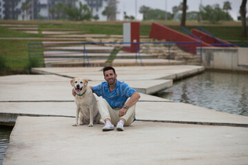 Handsome young man with beard and his Labrador retriever dog, posing for photos by a lake in the...