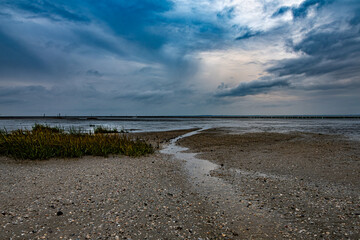 Ein Rinnsal fließt zwischen Muscheln und Wolken in Richtung Wattenmeer wo trotz Ebbe einiges an...