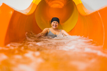 Laughing amused elderly caucasian female pensioner in black hair cap sliding on an orange water...