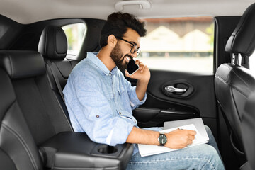 Smiling young indian businessman talking over phone, sitting in taxi