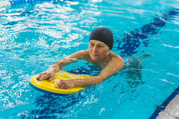 Elderly woman in black cap using yellow foam kickboard to swim in the indoor pool. High quality photo
