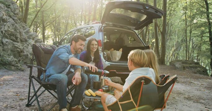 Father mother and children bonding in forest nature, drinking hot tea witch thermos sitting on table near parking car in sunny day. Attractive bearded man pouring hot drink from thermos to pretty kids