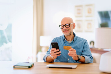 Confident mid aged man looking at smartphone while holding debit card in his hand at home