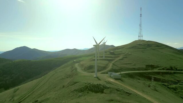 Wind turbines on the mountain against the blue sky. On the hills are trees and country roads, there is a communication tower. Renewable energy concept. Drone video. Climbing up. Pushkin Pass, Armenia.