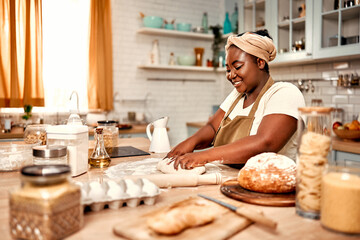 Baking at home. Side view of black housewife in traditional turban standing behind kitchen counter and working with dough and flour. Plus size woman with sincere smile on face crafting homemade bread.