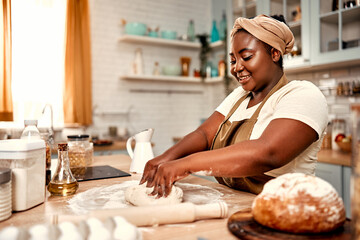 Homemade pastry. Curvy black woman kneading dough on kitchen table for creating foundation of pastries. Skillful housewife wearing brown apron and turban doing baked goods from traditional recipe.