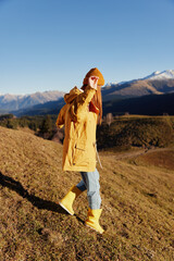 Woman back full-length walks on a hill and looks at the mountains in a yellow raincoat and jeans in the autumn happy trip in the sunset sunset hiking, freedom lifestyle 