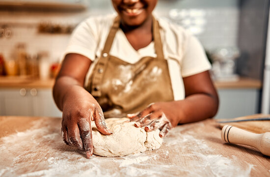 Family recipe. Close up of smiling curvy woman in brown apron shaping raw dough in flour on wooden kitchen table. African american lady preparing traditional homemade bread during daily routine.