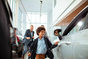 Excited young boy exploring a car at a dealership with his father watching