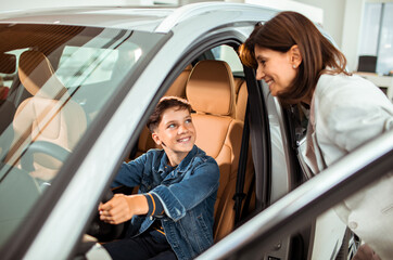 Excited boy sitting in a car while his mother looks on at a dealership