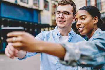 Positive dark skinned hipster girl with caucasian friend making photo on phone for blog