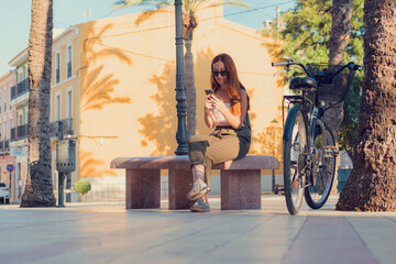 Young woman sitting on a bench with her bicycle looking at her phone in a town