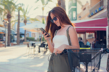 Young woman with a bicycle looks at her phone in a town at sunset