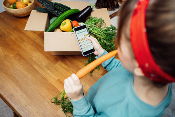 Woman holding phone with active online mobile app of Veganuary diet calendar and eating fresh carrot above box with vegetables. Healthy eating, weight loss. Food delivery, recipe box. Selective focus.