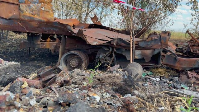 An unexploded mine lies on side of road against the background of rusty car blown up by mine among planting of trees. War in Ukraine