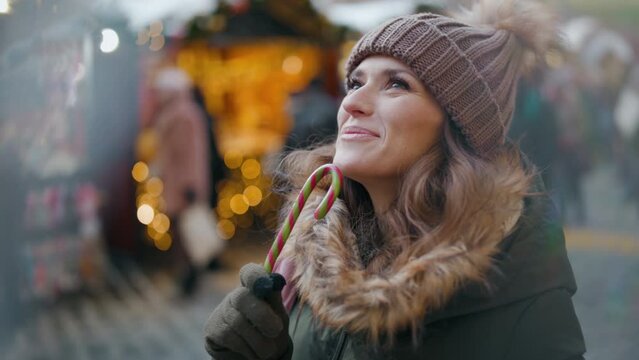 smiling modern woman in green coat and brown hat at the winter fair in the city with candy cane.