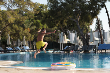 Young boy joyfully jumping into a clear blue swimming pool with a colorful inflatable ring,...