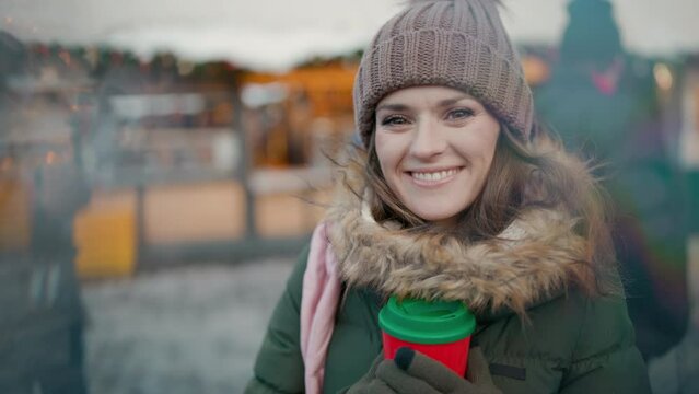 happy young female in green coat and brown hat at the christmas fair in the city with red cup of hot Christmas beverage.