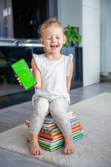 Happy Little girl is sitting on stack of children's books and holding a smartphone with a chromakey
