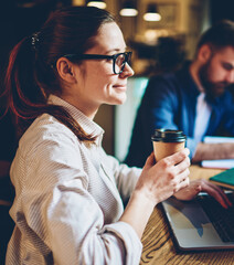 Side view of prosperous female student in spectacles holding tasty coffee in hands 
