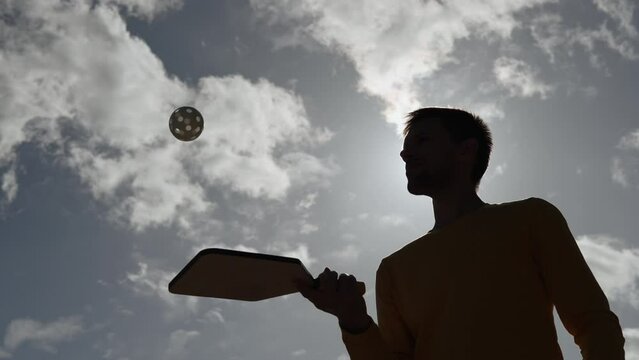 Silhouette of man deftly hitting pickleball ball with racket against sky. Player practicing hitting plastic ball with round holes during training outdoors in sunny weather.