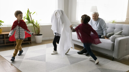 Fototapeta premium Aged great-grandfather spend time with three great-grandsons at home, kids in festive Halloween costumes running in living room, having fun together, enjoy active game, friendship and family pastime