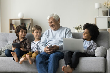 Loving older great-grandpa and great-grandsons sit on couch holding diverse modern devices, laugh,...