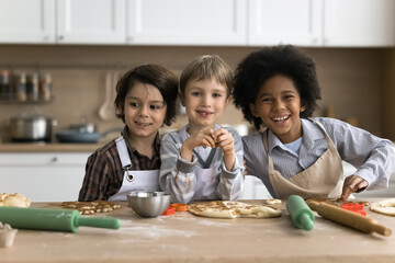 Three multiethnic preschoolers boys in aprons cooking together in kitchen, prepare surprise for family, make dough for homemade pizza posing smile look at camera at home. Cousins friendship and hobby - obrazy, fototapety, plakaty
