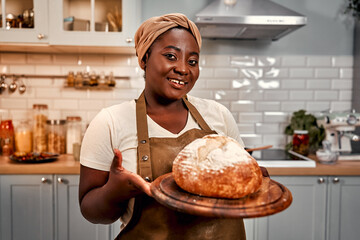 Handmade bakery. Happy hostess in headband and apron holding freshly baked crunchy bread on wooden board. Smiling curvy woman with black skin proudly looking at camera while standing on home kitchen.