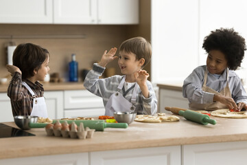 Three cute multiethnic little boys wear aprons give high five, cooking pizza, Italian cuisine gathered together in modern domestic kitchen, sit at table enjoy communication and cookery. Family, hobby