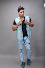 Indian young male model with short black hair in casual wear wearing black t-shirt, blue denim jacket and blue torn denim jeans, posing against gray background. Fashion Portrait, Advertisement shot.