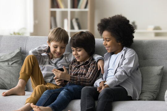 Three smiling cute boys sit on sofa in living room use mobile phone play online games on smartphone, watch funny video, friends spend free time together at home chatting with mates. Modern technology