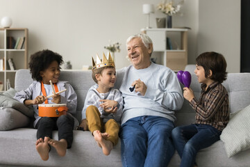 Great-grandpa having fun with three little great-grandchildren, sit together on sofa and play...