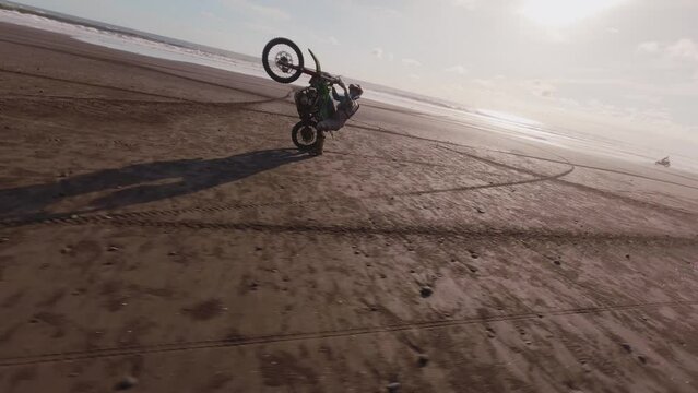 Professional sportsman enduro motorcycle driving sandy tropical beach performing extreme tricks riding on one back wheel. Man biker enjoying dangerous riding wheely bike nature. Aerial FPV drone view