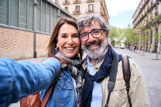 Selfie portrait smiling mature Caucasian tourist couple posing outdoors. Grizzled man and adult woman affectionate embracing. Happy people in love in sixties looking at camera together enjoying city.