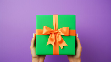 Anonymous person showing bright green carton gift box with ribbon and purple bow on orange table against violet background - obrazy, fototapety, plakaty
