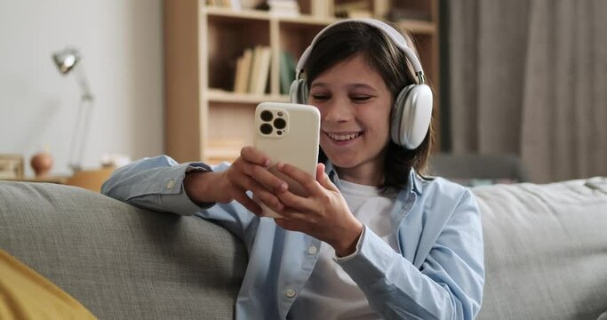 Schoolboy wearing headphones laughs heartily while using phone on the couch. His infectious laughter and enjoyment of digital content create a scene filled with youthful exuberance.
