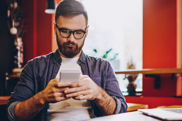 Serious male blogger in eyewear for vision correction reading sms with notification for app on smartphone