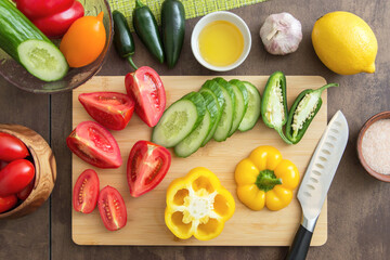 Different sliced vegetables top view for cooking salad on cutting board with knife on table on kitchen. Avocado, pepper, tomato, cucumber, oil, salt