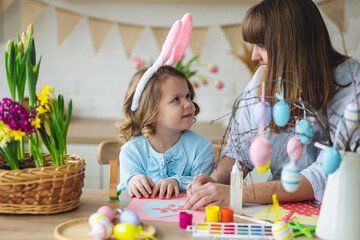 Mother and daughter painting eggs, creating home Easter decor together, happy, cheerful. Cute pretty smiling little girl, family time. Fresh spring bright colourful flowers in the kitchen. Sunny day